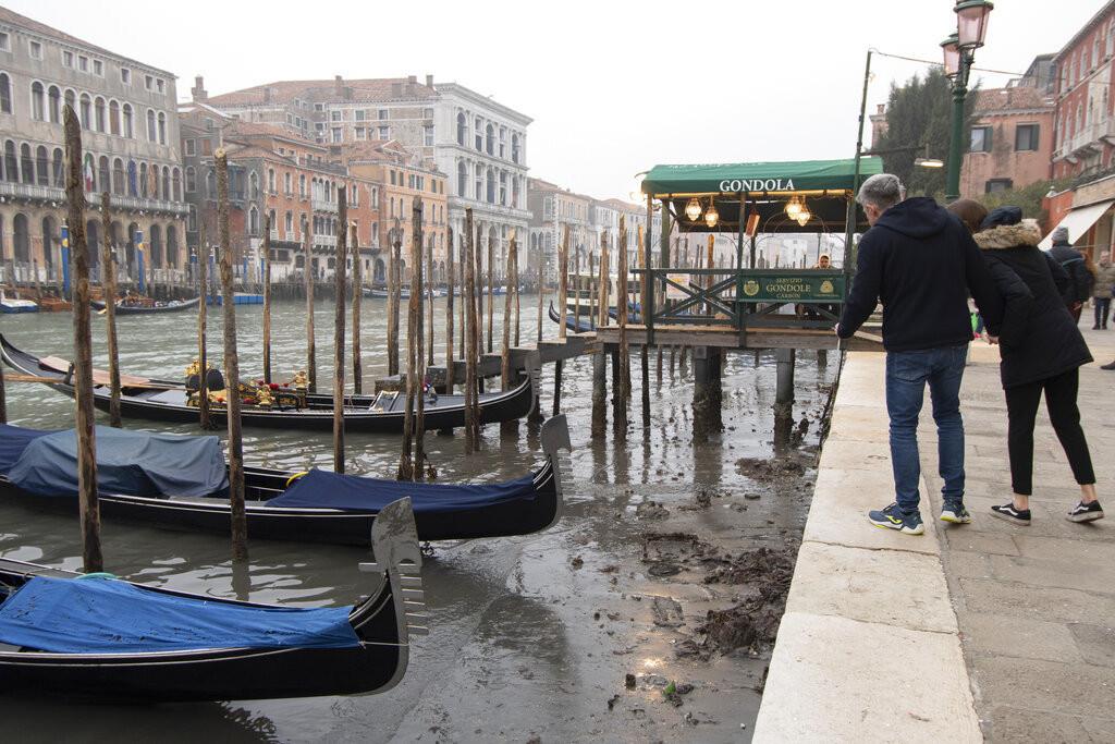 Varias personas observan las góndolas atracadas en un canal durante la marea baja en Venecia, Italia, el martes 21 de febrero de 2023. (AP Foto/Luigi Costantini)