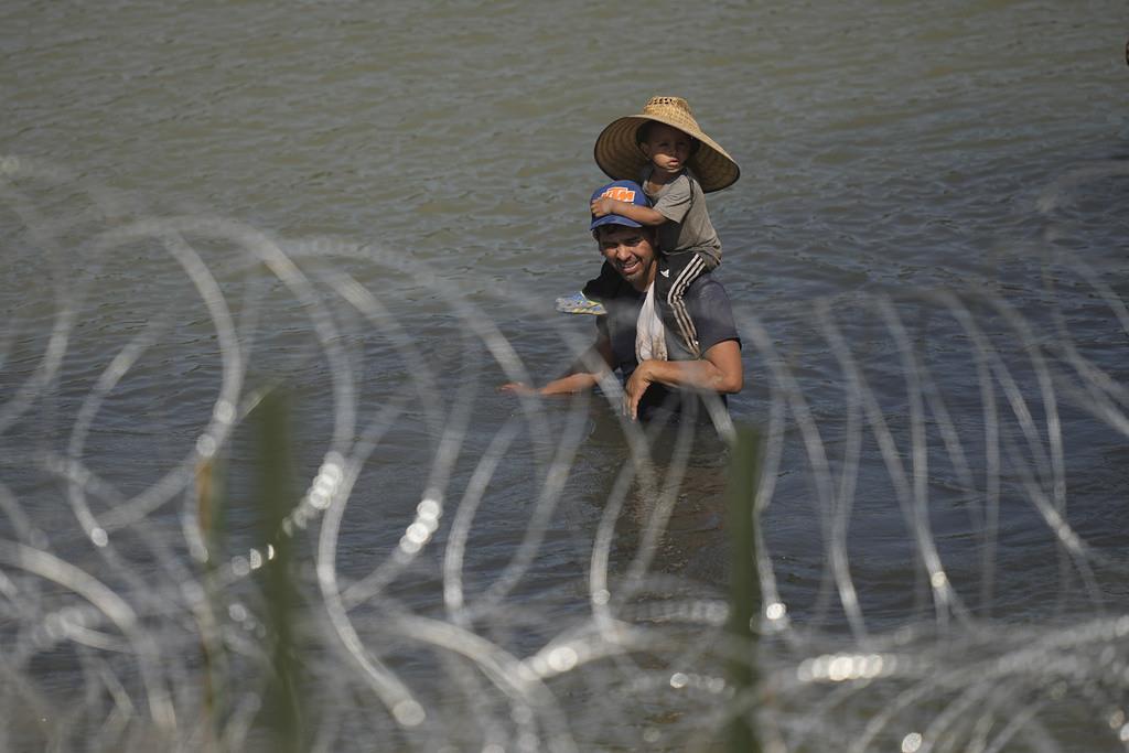 Un migrante cruza el río Bravo con un niño sobre sus hombros en un intento por llegar a Estados Unidos desde México, cerca del lugar donde trabajadores instalan una fila de boyas para que sean una barrera flotante, el martes 11 de julio de 2023, en Eagle P