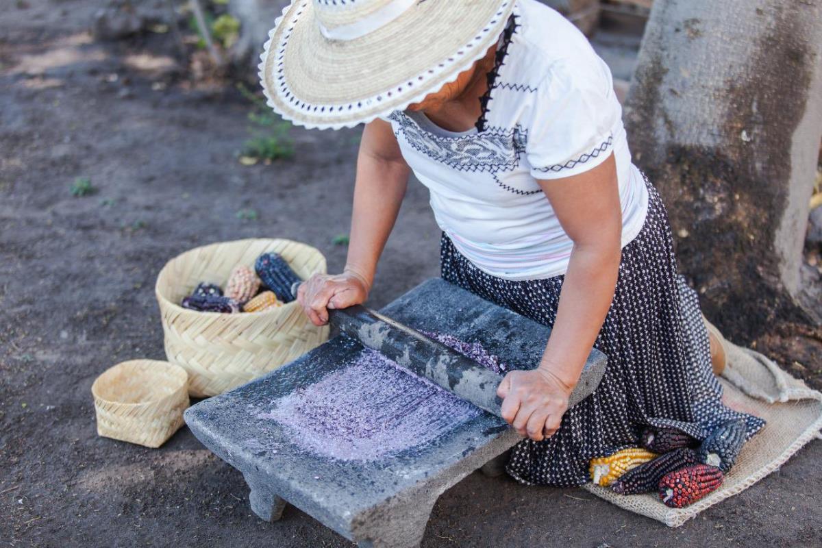 Mujer muele maíz en un metate. Créditos: X (@SECTUR_mx).