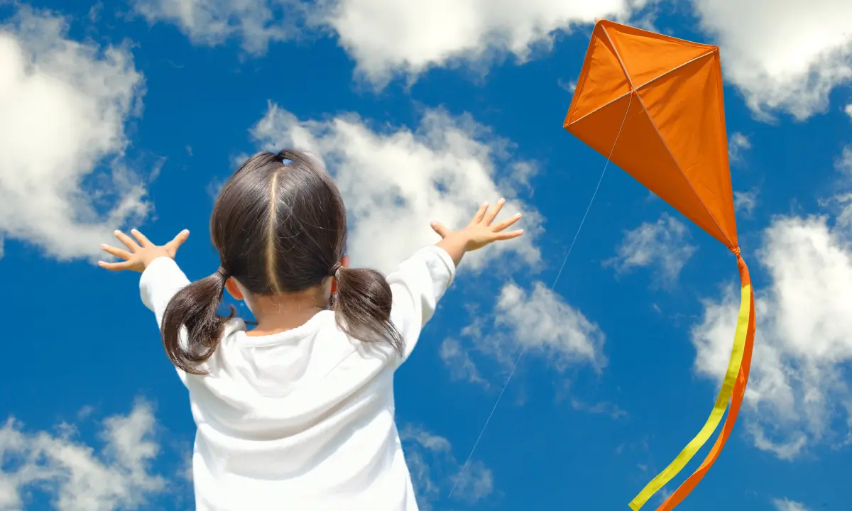 Niña de espaldas con los brazos al aire y un papalote naranja en el cielo Foto: ChatGPT/Canva