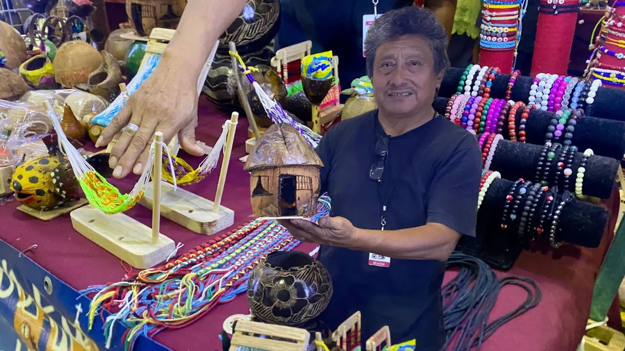 Al fondo, parte de las artesanías que se ofrecen en el marco de la feria Yucatán Xmatkuil 2025. Al frente, uno de los artesanos muestra sus productos en la nave del IYEM.- Fuente Feria Yucatán